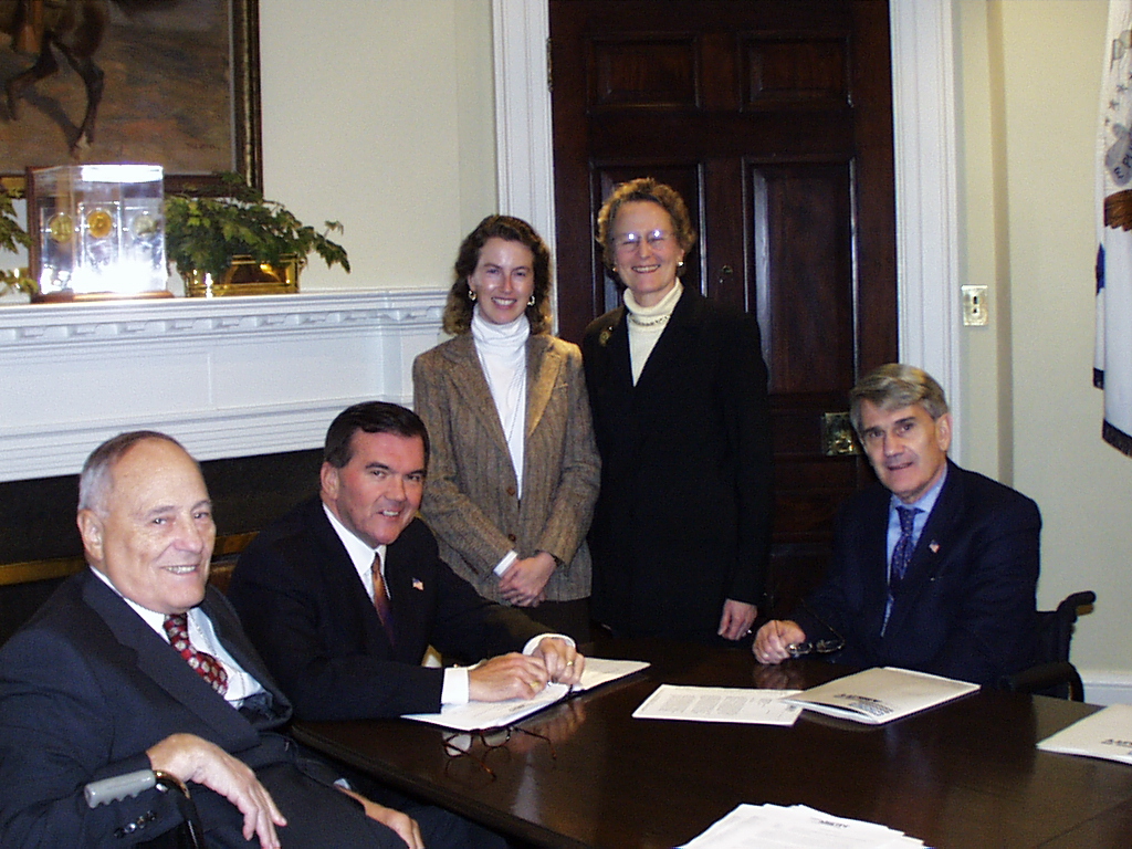 Photo caption: Alan Reich, graying hair in a white collared shirt and dark suit and tie in a wheelchair. Seated next to him is former PA Governor Tom Ridge, then Director of the Department of Homeland Security, with black hair, a white collared shirt and a dark suit.  Standing is me – Mary Dolan – with a white turtleneck, brown jacket, and brown shoulder-length hair. Next to me is Ginny Thornburgh, in a white turtleneck and black jacket and grey hair. To the right is Mike Deland, seated in a wheelchair, in a dark suit, white collared shirt and tie.